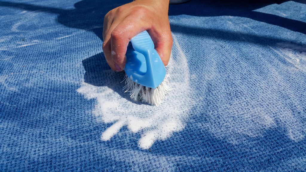 Teppichreinigung - Eine Hand, welche einen Teppich mit einer Bürste sauber macht.